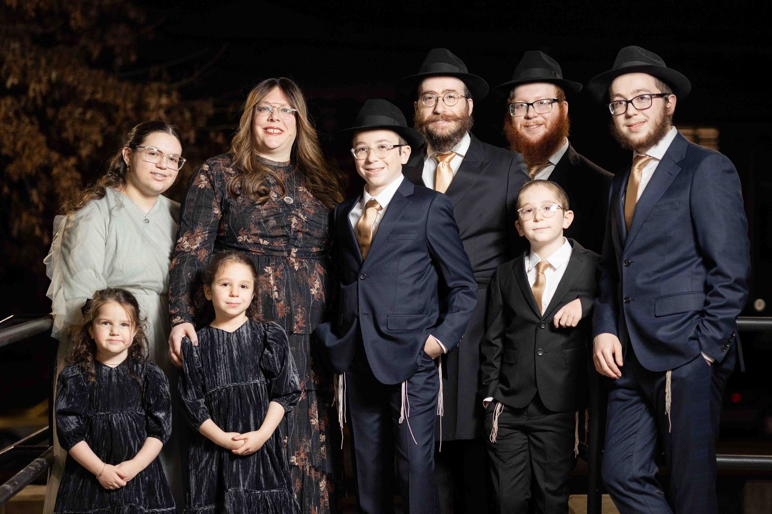 Large Jewish family poses together outdoors at night in formal attire for a special occasion.