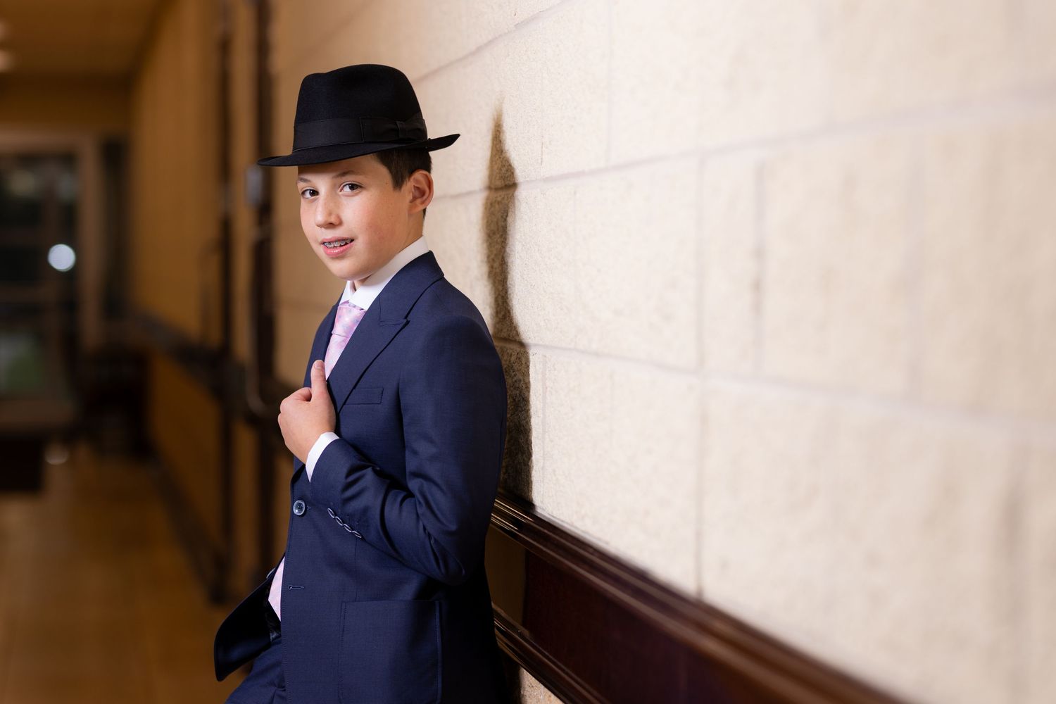 Young man in navy suit and black fedora hat posing confidently against a beige stone wall in a hallway.