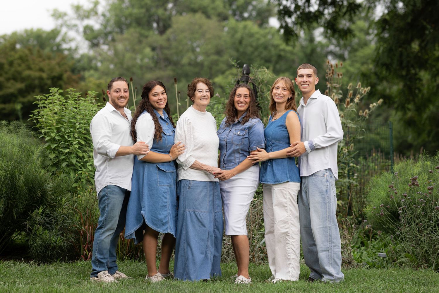 A smiling family of six poses outdoors in a lush green garden, wearing coordinated blue and white outfits.