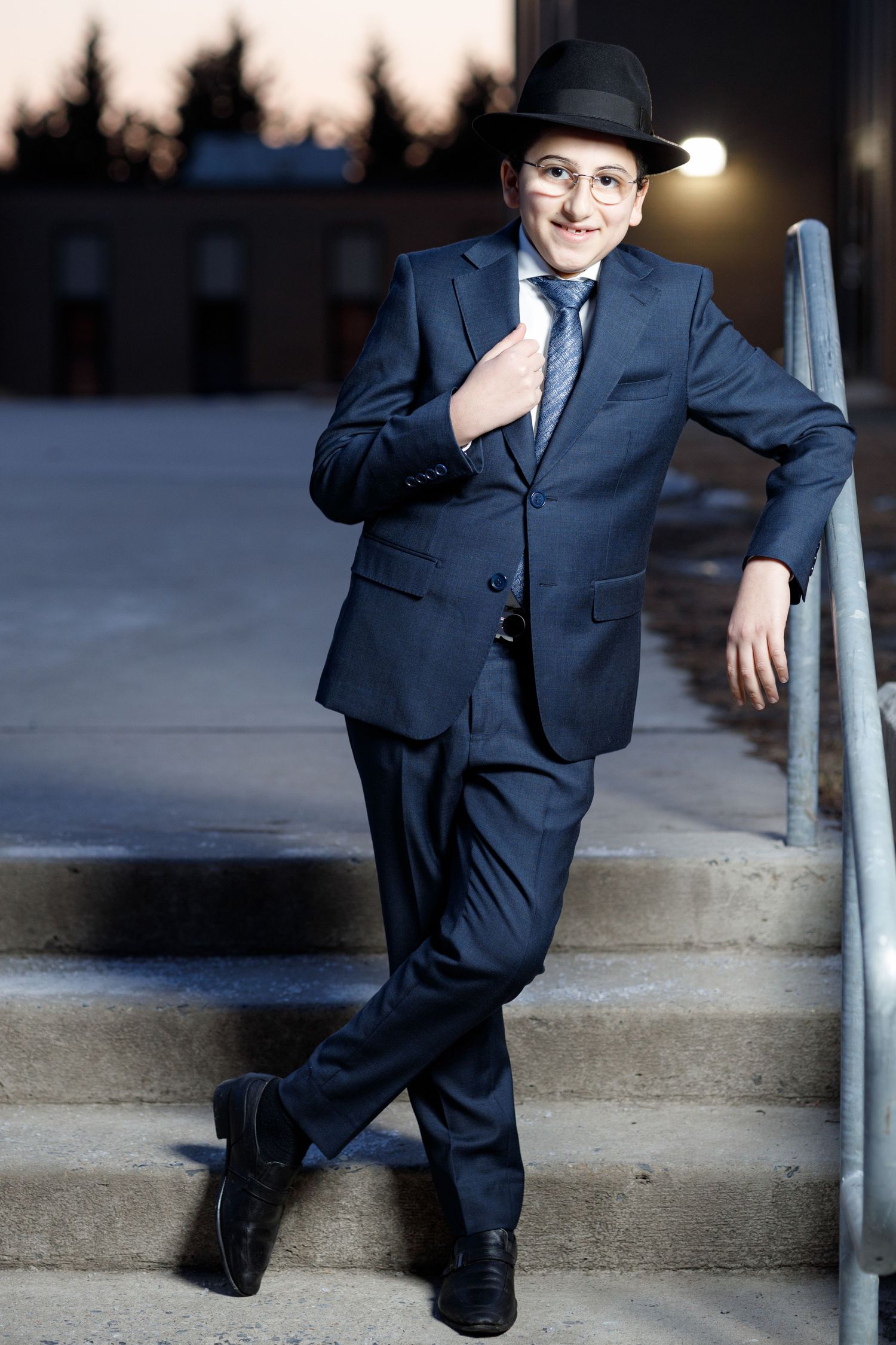 A young boy in a navy suit and fedora poses confidently on outdoor steps at dusk.