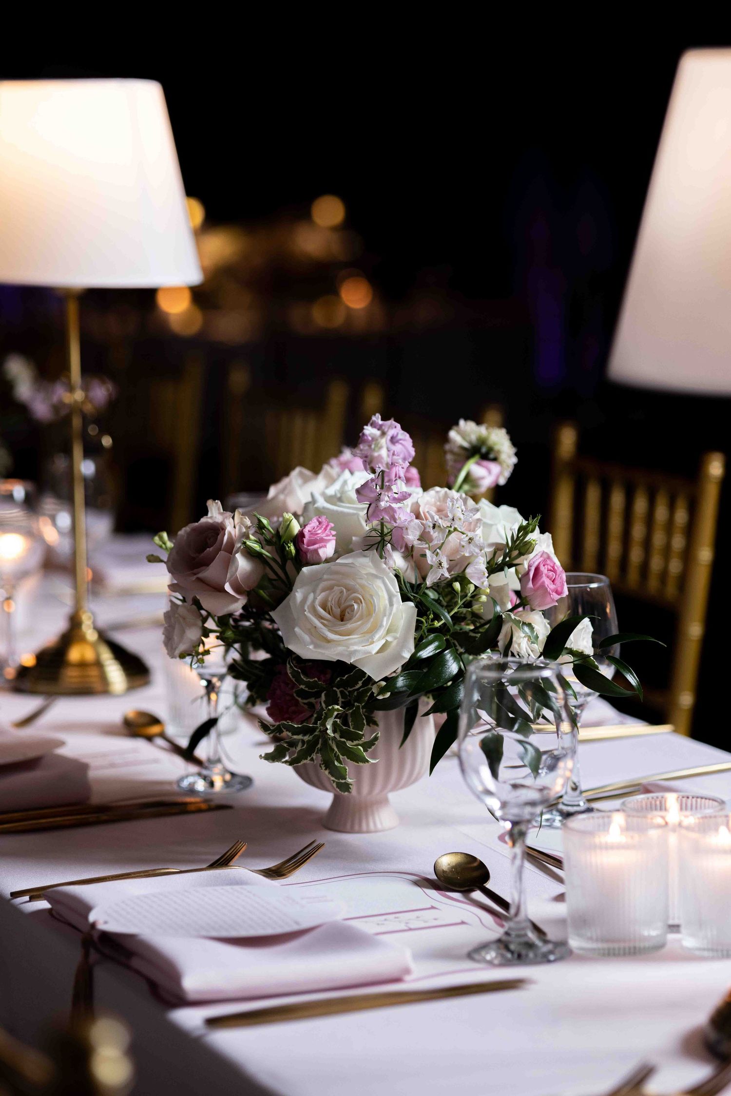 Elegant wedding table setting with white and pink floral centerpiece, gold cutlery, candles, and white table lamps at night.