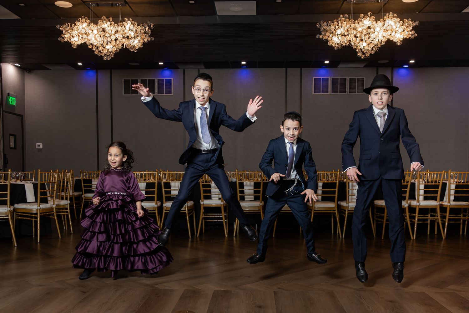 Four people in formal attire joyfully jumping in an elegant banquet hall with chandeliers.