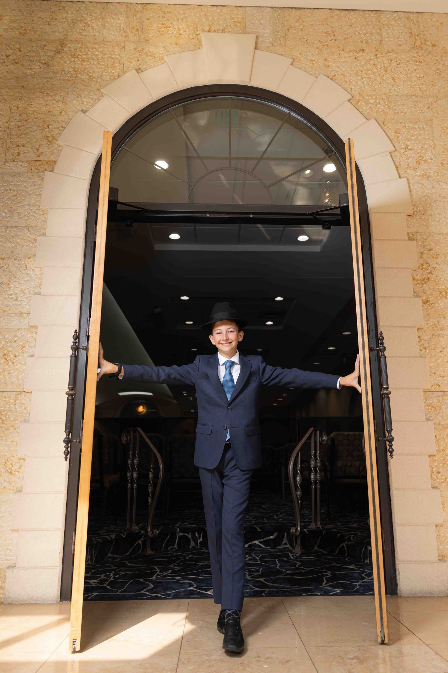 Young boy in blue suit and black hat stands in elegant arched doorway with arms outstretched.