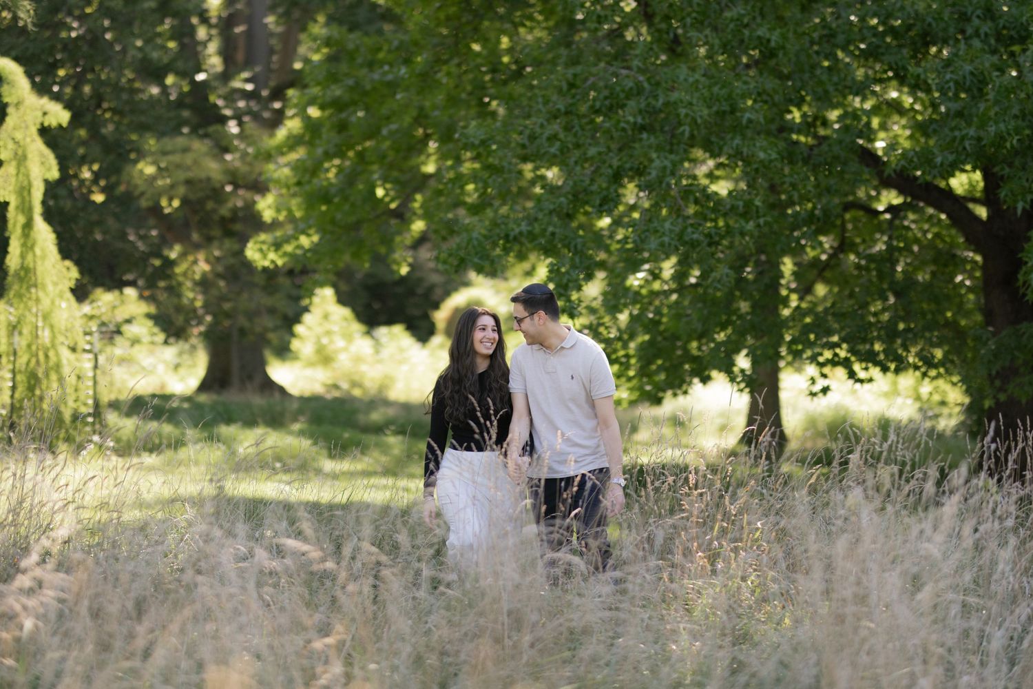 Couple walking hand in hand through tall grass in a lush green park surrounded by trees.