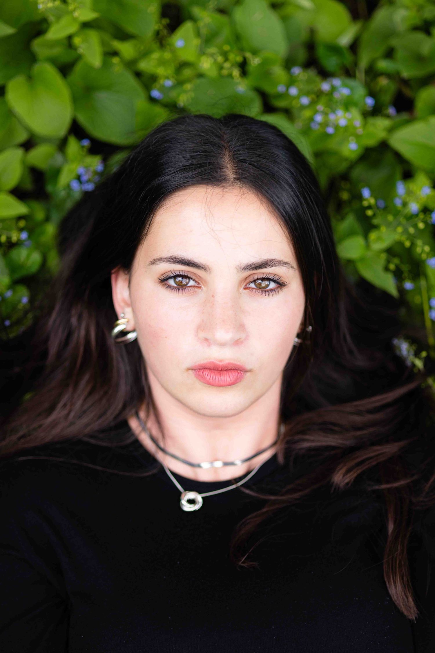 Close-up portrait of young woman with dark hair lying among green foliage and blue flowers.