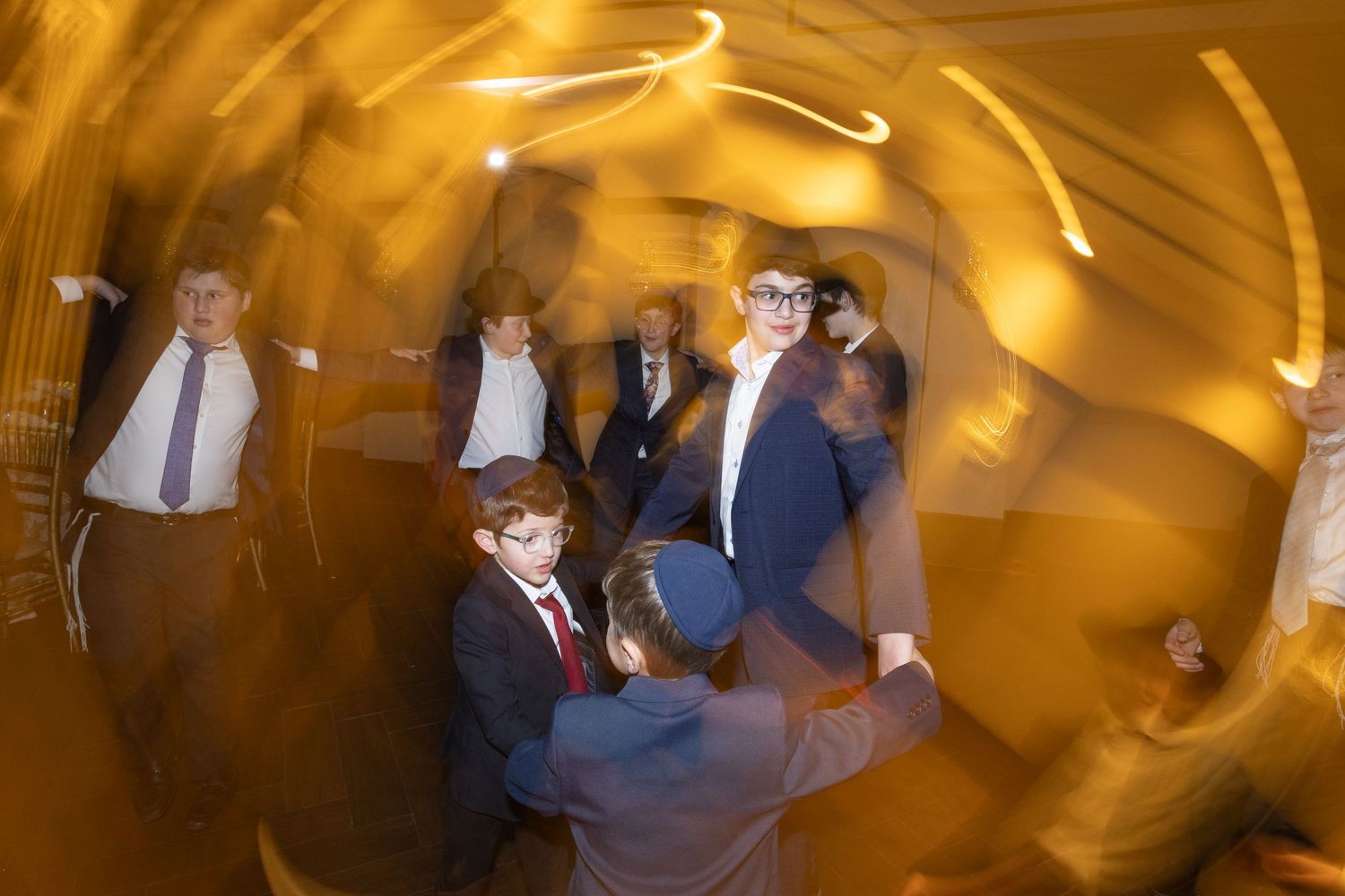 Men and boys in suits dance joyfully at Bar Mitzvah with swirling golden light effect.
