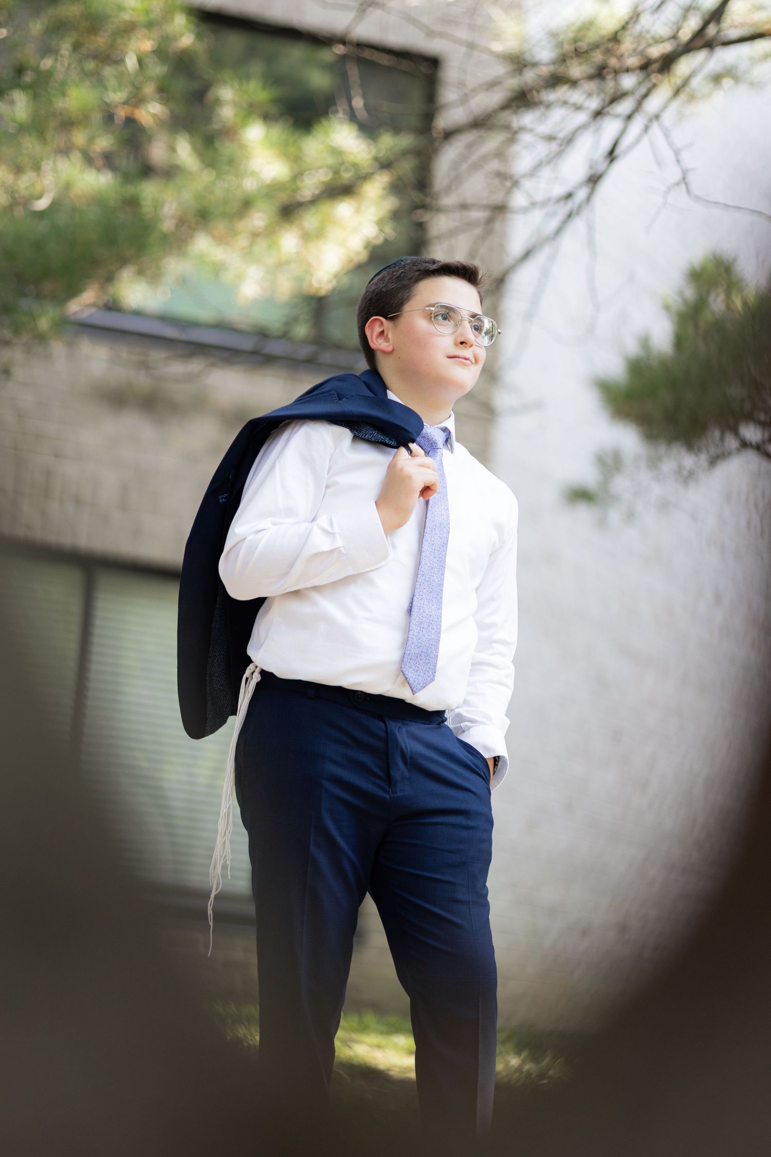 A young boy in a white shirt and blue tie carries his jacket over his shoulder outdoors.
