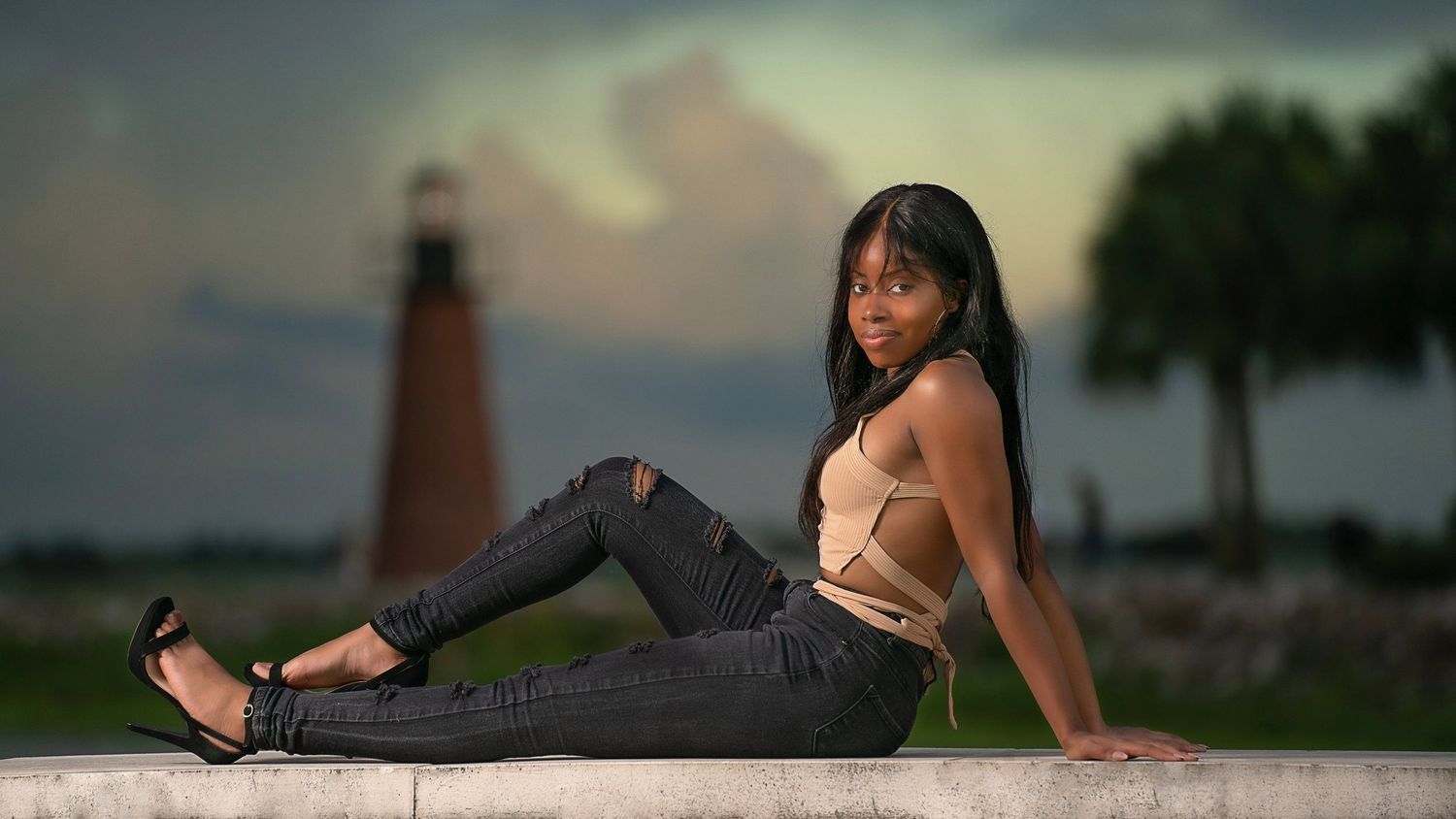 Young woman in ripped jeans poses near a lighthouse at dusk.