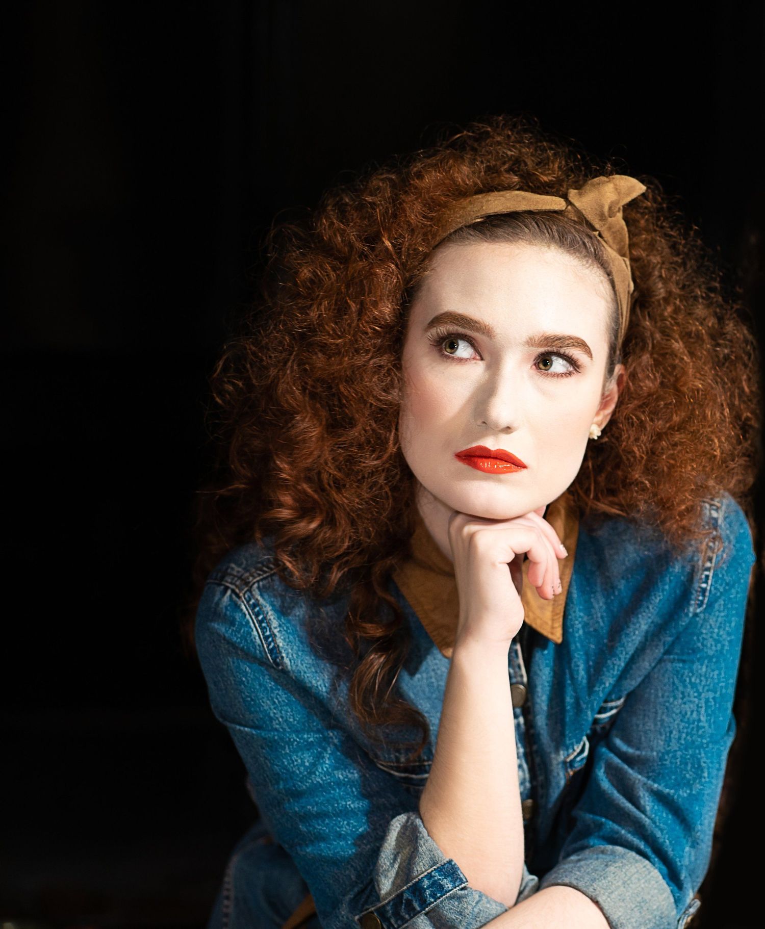 Young woman with curly red hair and red lips, wearing denim shirt and gold headband, posing thoughtfully against dark background.