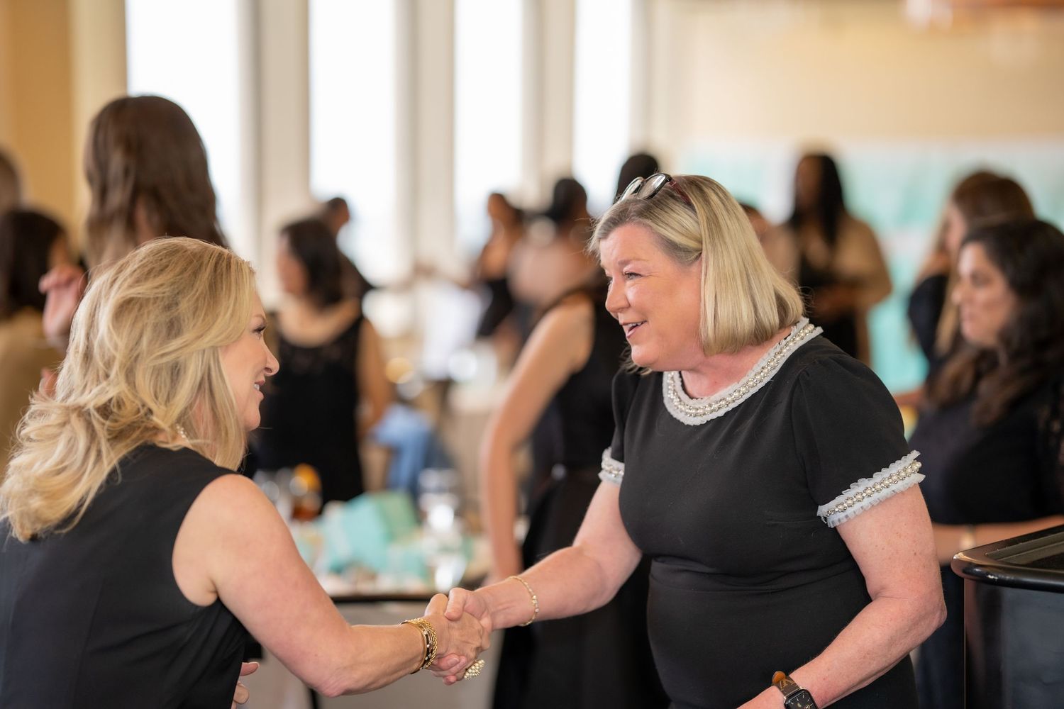 Two women in black dresses shaking hands and smiling at a networking event with guests in background.
