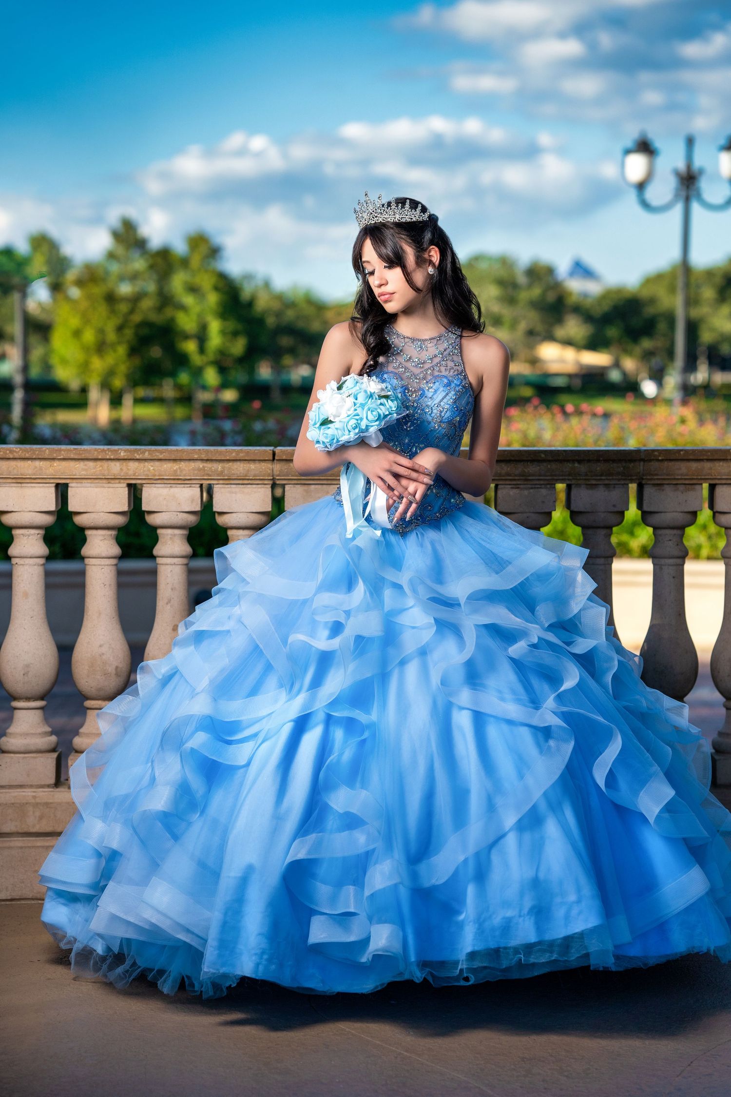 Young woman in a blue ball gown with tiara holds bouquet at outdoor balustrade for quinceañera.