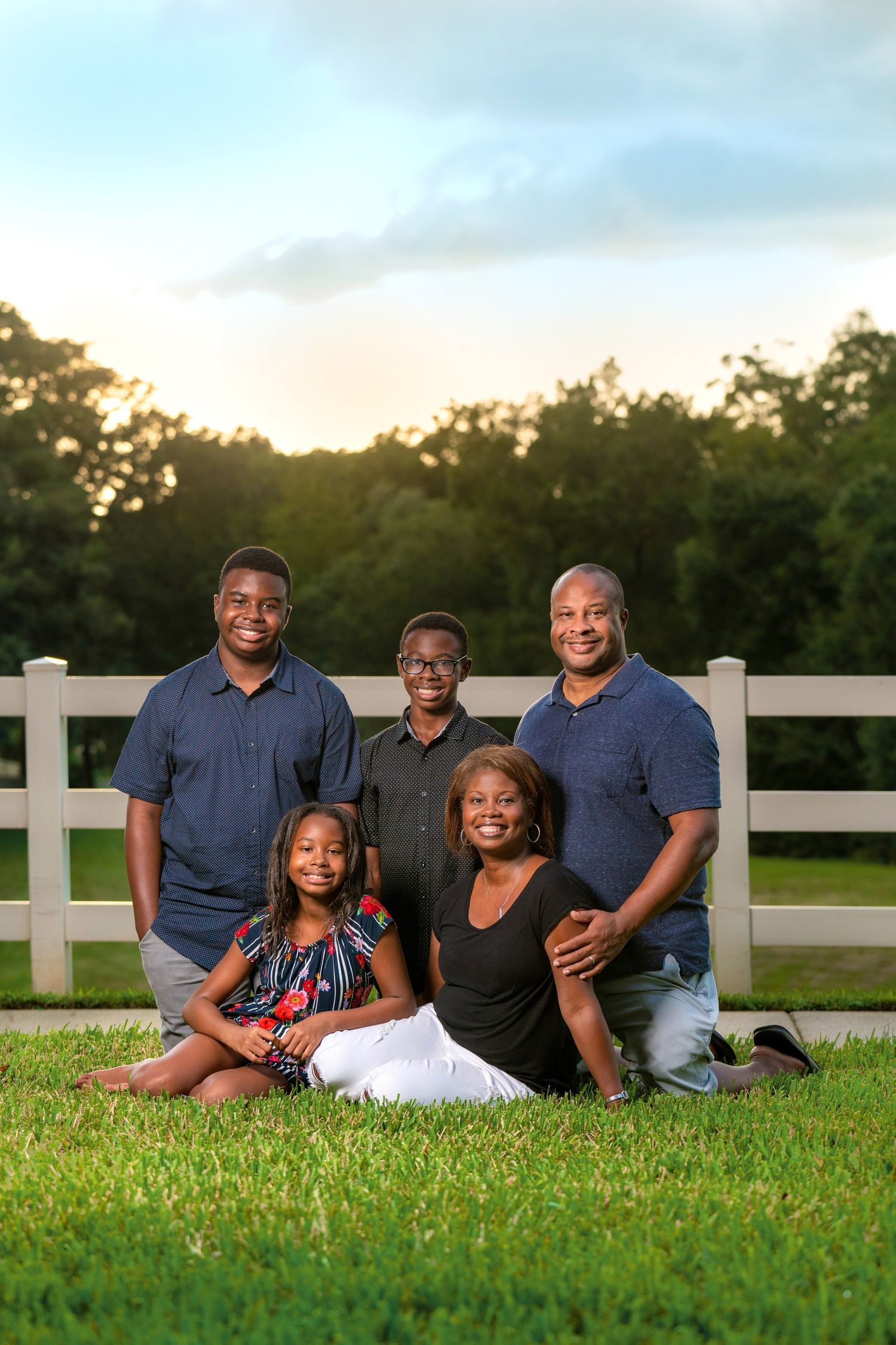 A family of five poses together on grass in front of a white fence during sunset.