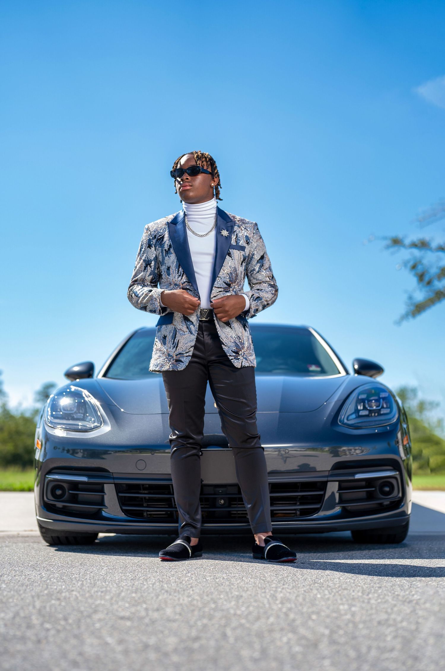 Stylish man in floral blazer and sunglasses posing confidently in front of a gray Porsche on a sunny day.