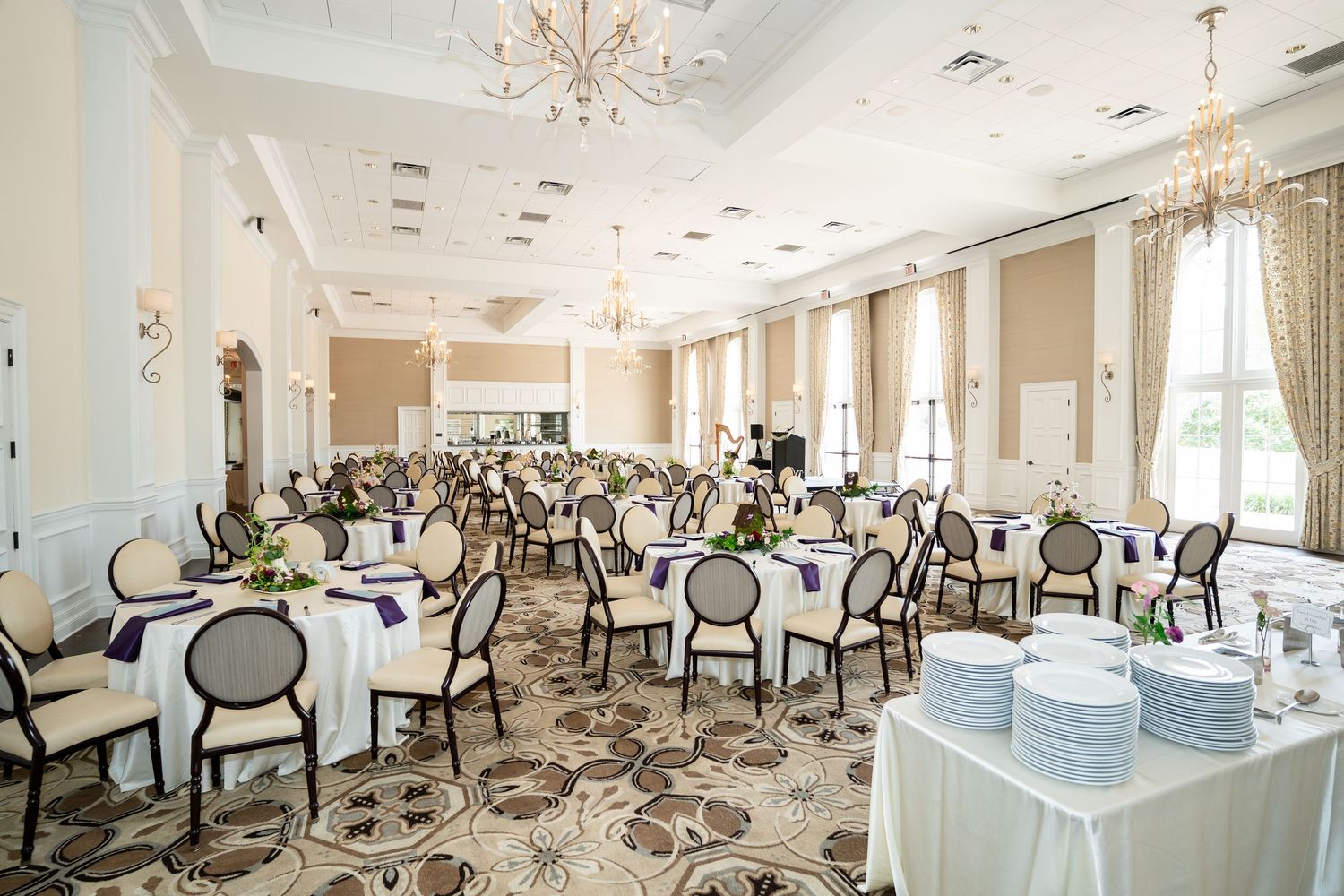 Elegant ballroom with white-draped tables, purple accents, chandeliers, and ornate carpeting for a formal event.