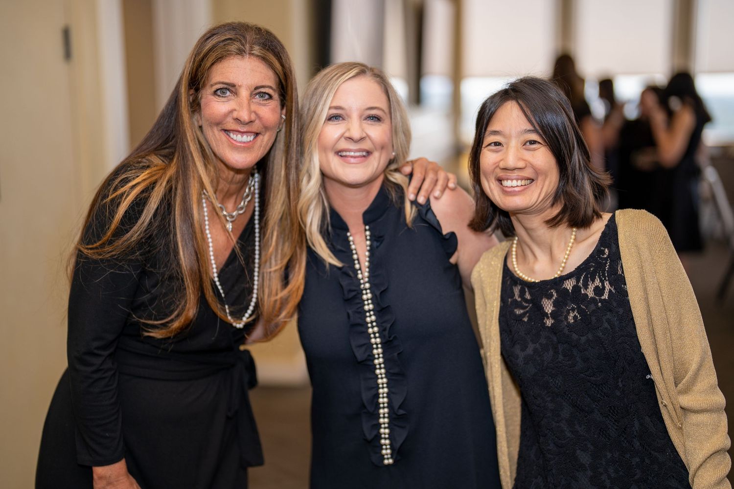 Three women smiling and posing together at a formal event, wearing elegant dark outfits in a bright indoor venue.