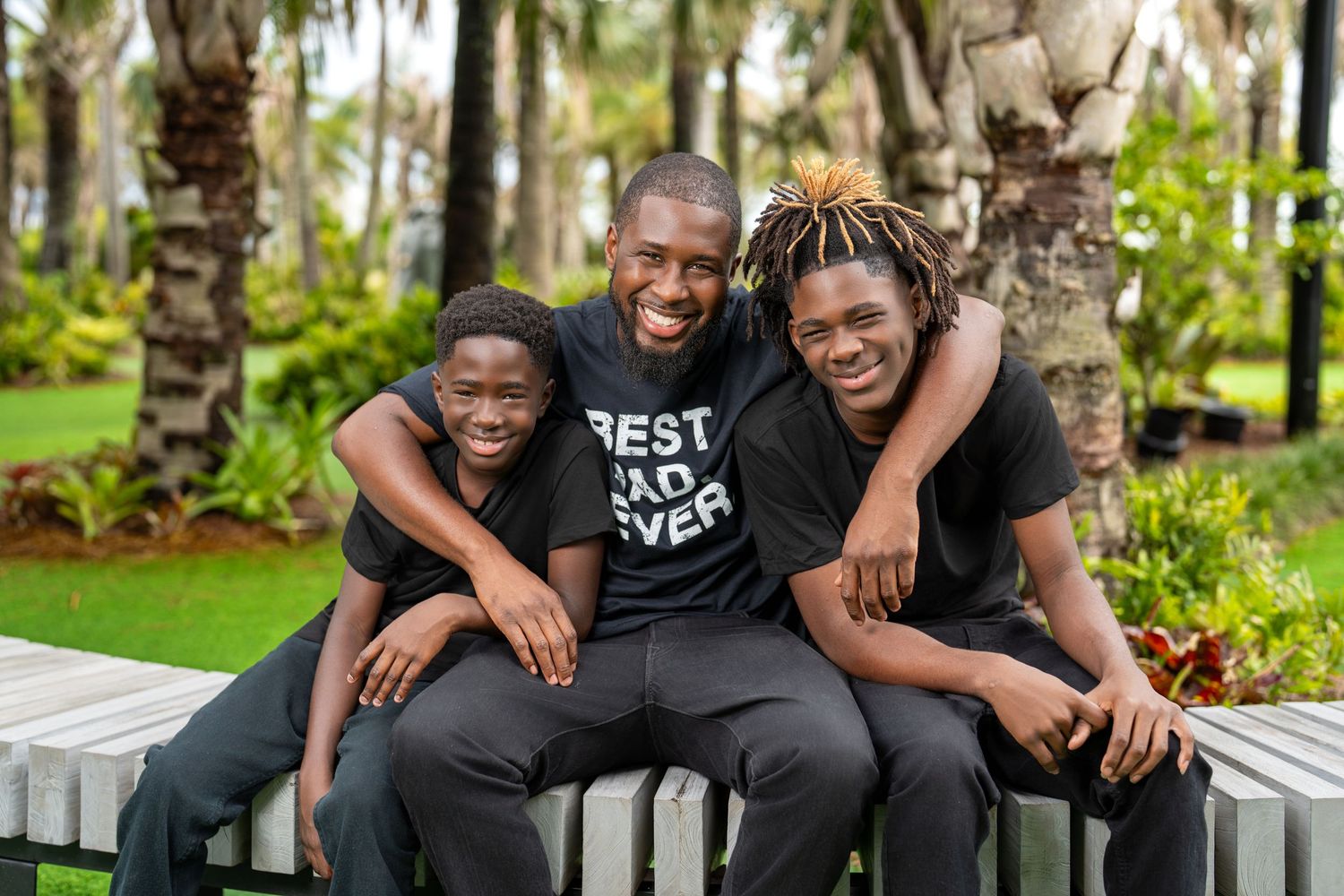 A father wearing a Best Dad Ever shirt smiles with his two sons on a bench surrounded by palm trees.