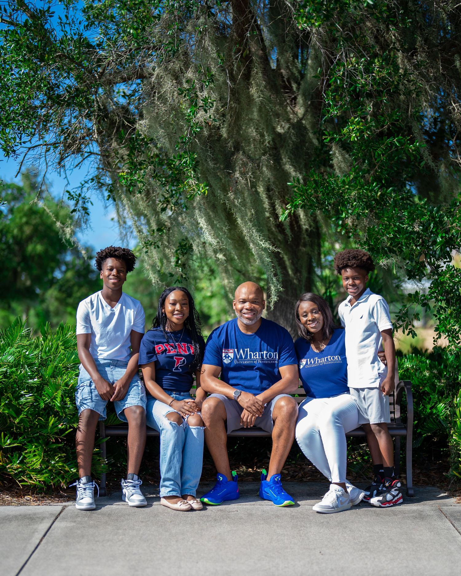 Family of five smiling together on a park bench surrounded by lush greenery, wearing college-branded shirts outdoors.