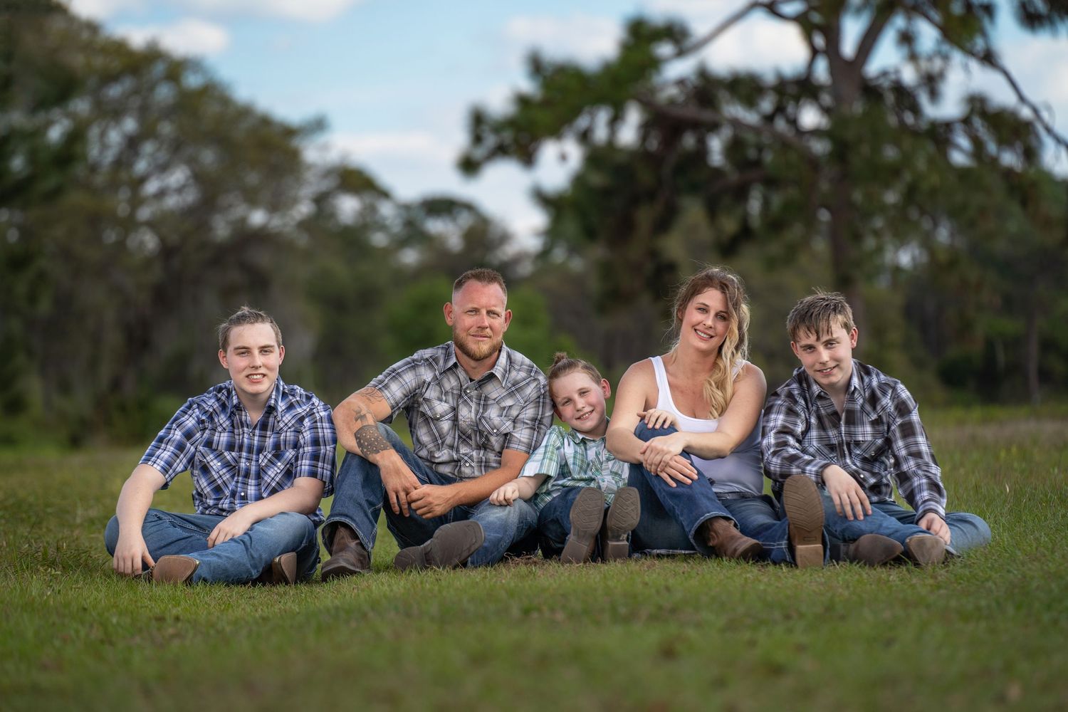 Family of six sitting on green grass outdoors, wearing matching plaid shirts, smiling together in a park setting.