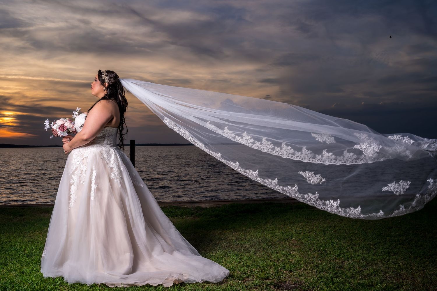 Bride in white gown with flowing lace veil blowing in wind at waterfront sunset backdrop.