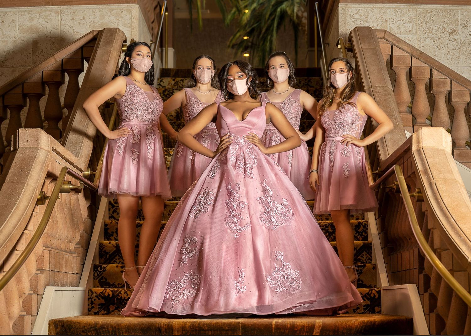 Five young women in pink dresses and masks pose on ornate stairs during a quinceañera celebration.