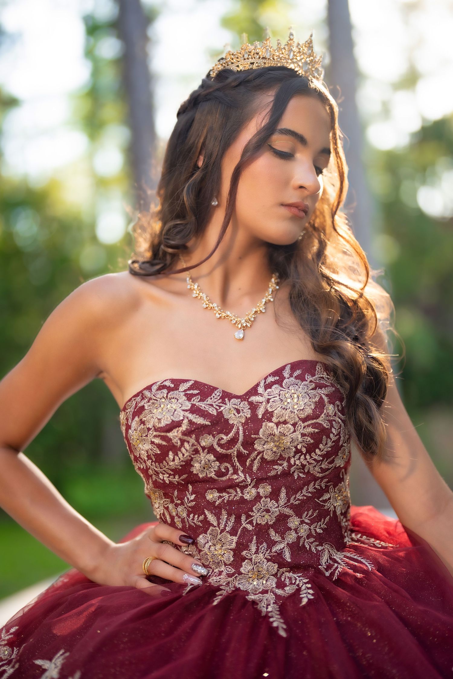 Young woman in burgundy quinceañera gown with gold embroidery, crown, and necklace posing outdoors.