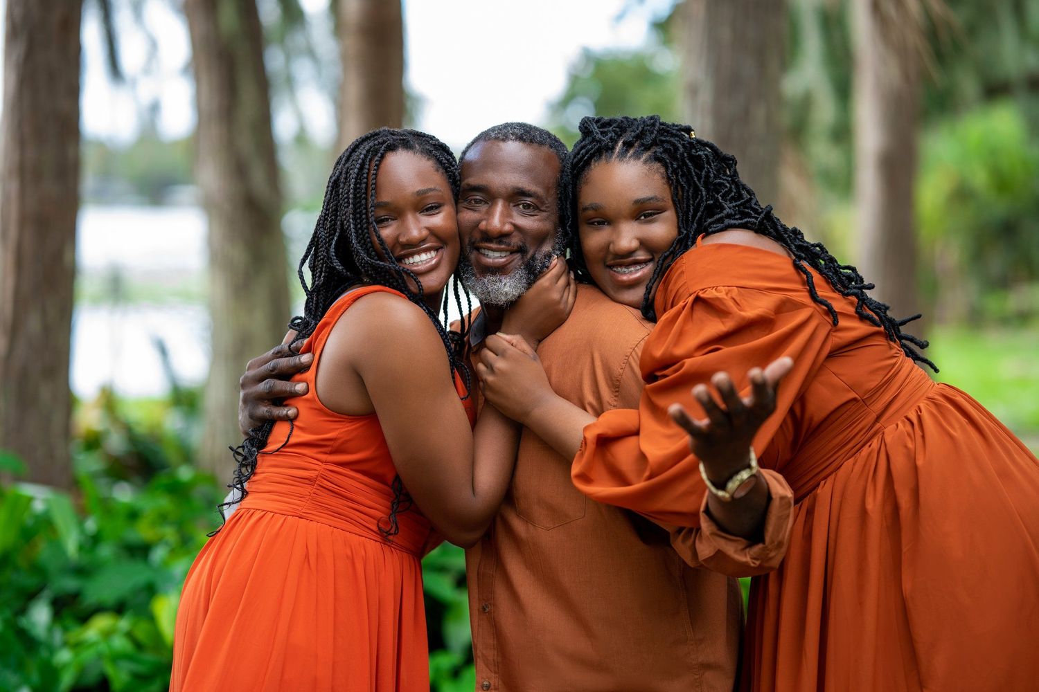 Three people in bright orange dresses embrace warmly outdoors.