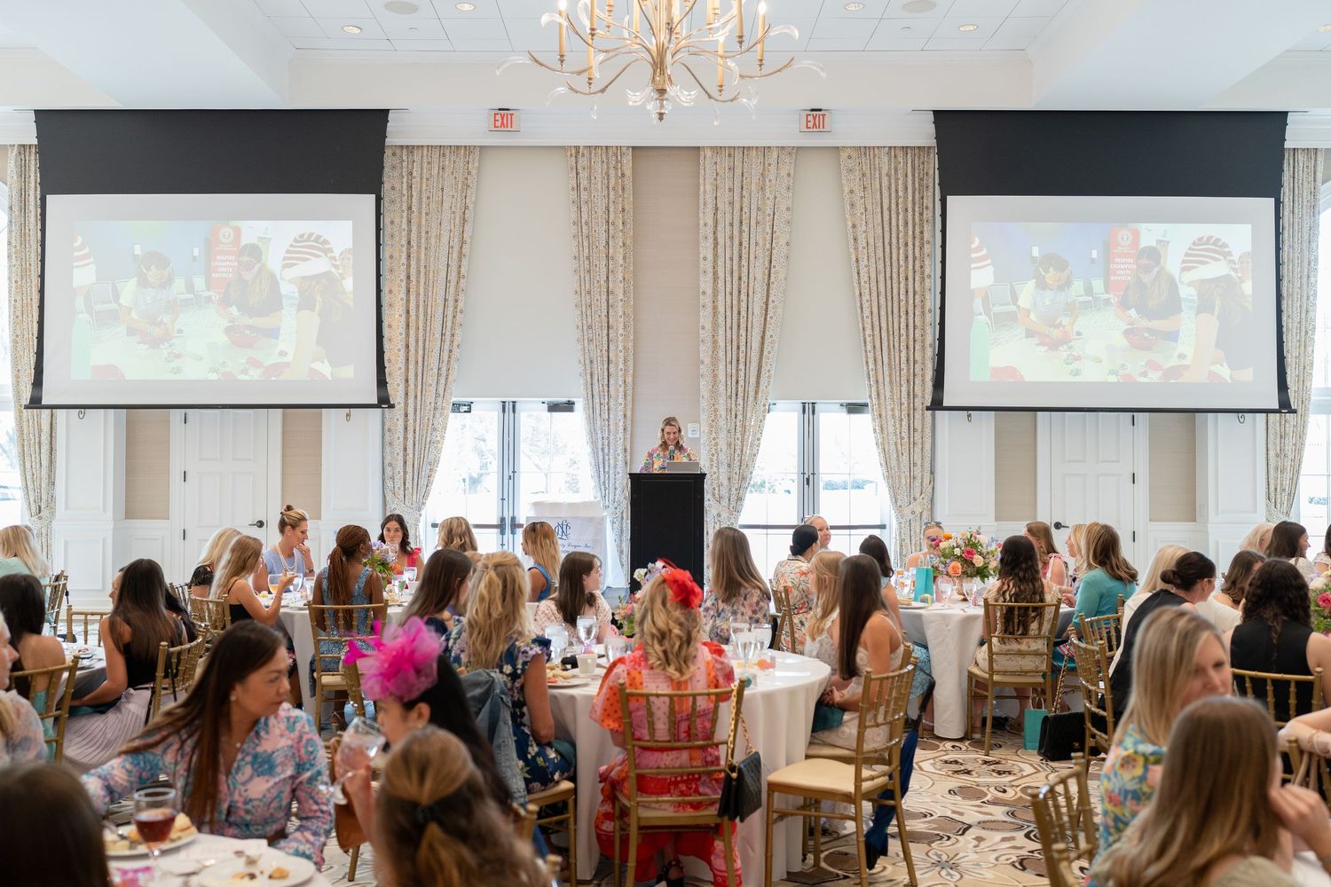 Elegant women's luncheon event in a ballroom with projection screens, chandelier, and colorful floral arrangements.