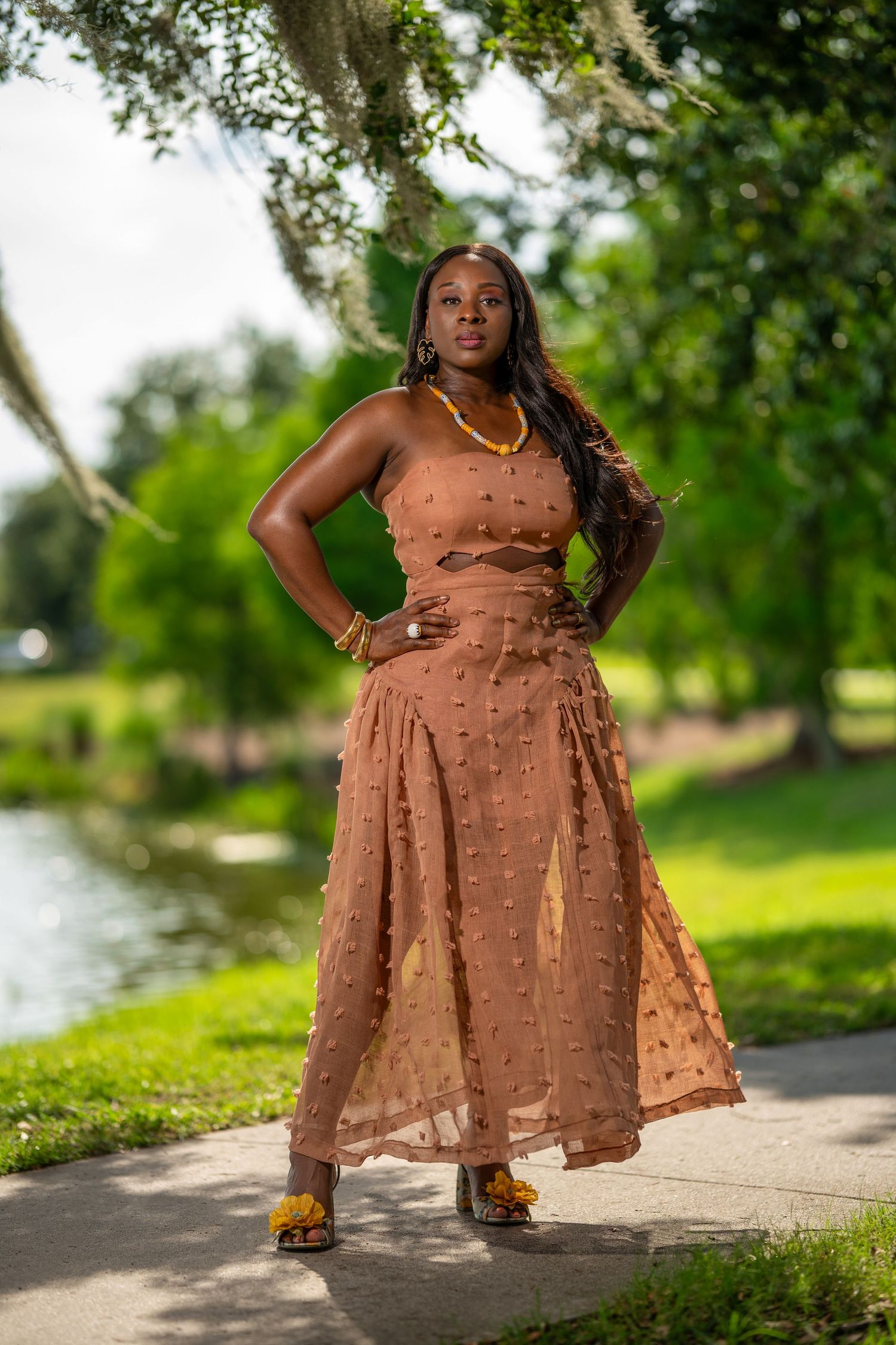 A woman in a brown textured midi dress poses confidently outdoors near a pond in a lush green park.