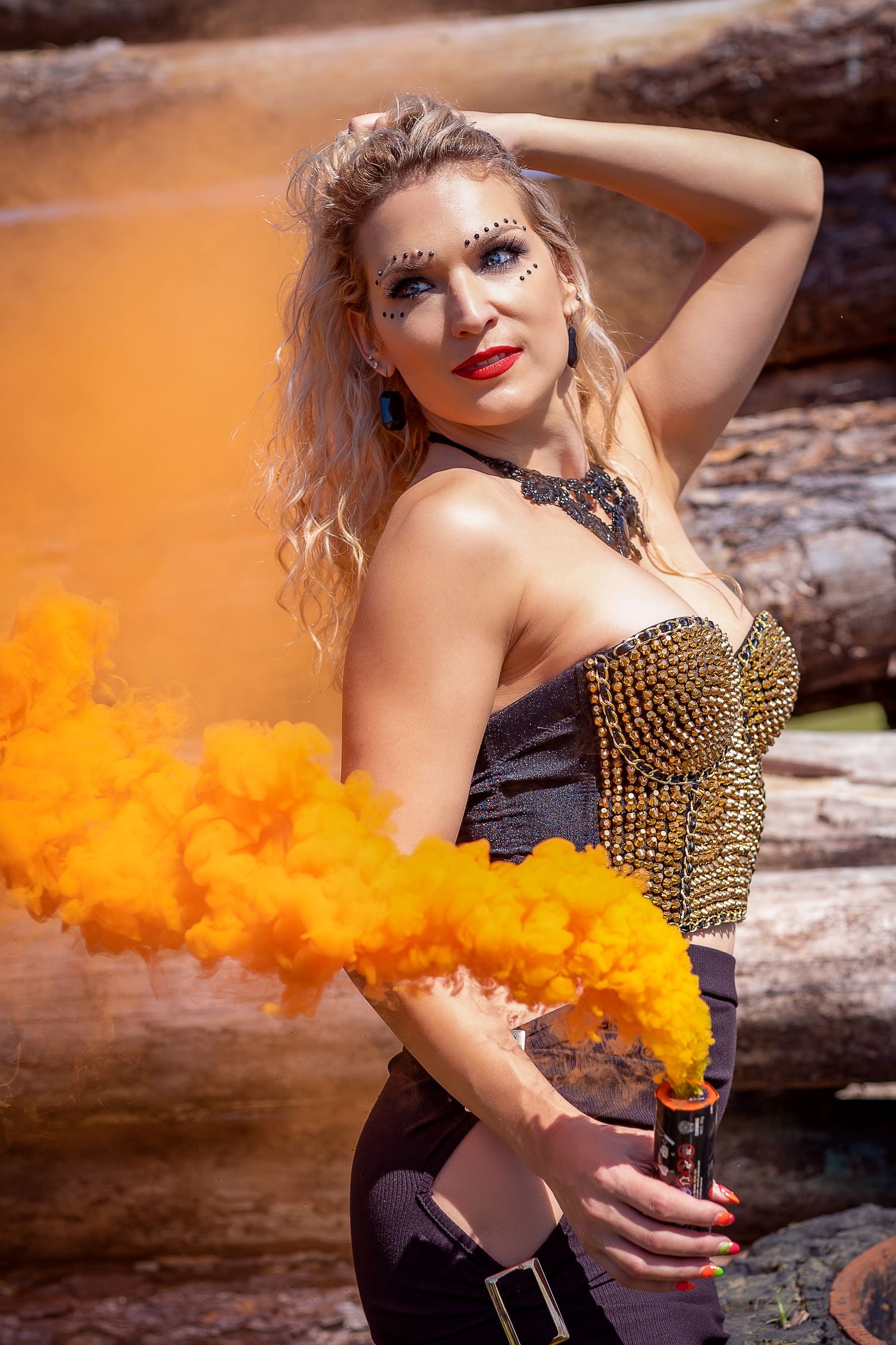 Woman in studded corset poses with orange smoke flares against rustic backdrop.