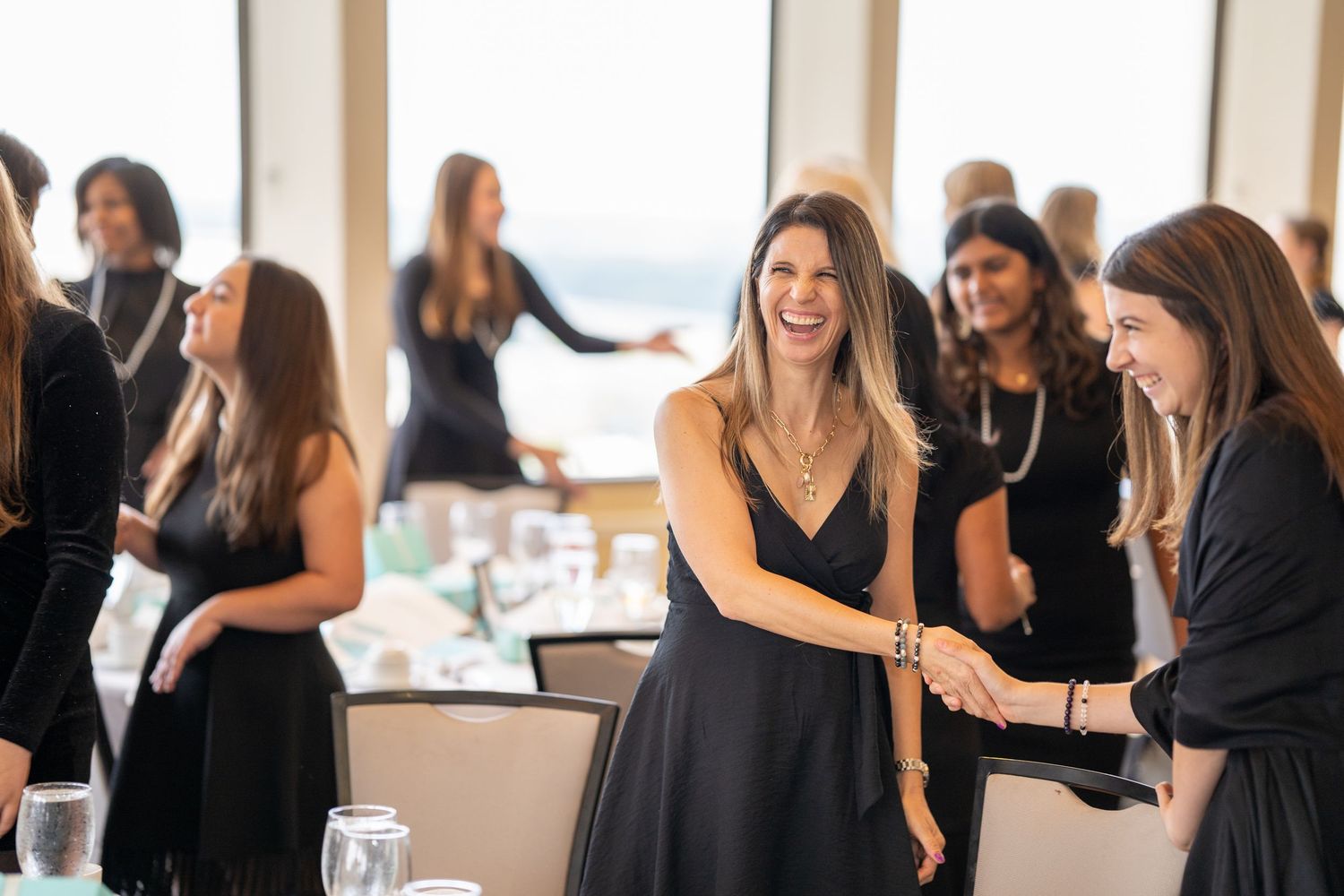 Women in black dresses networking and laughing at an elegant indoor event with ocean views.