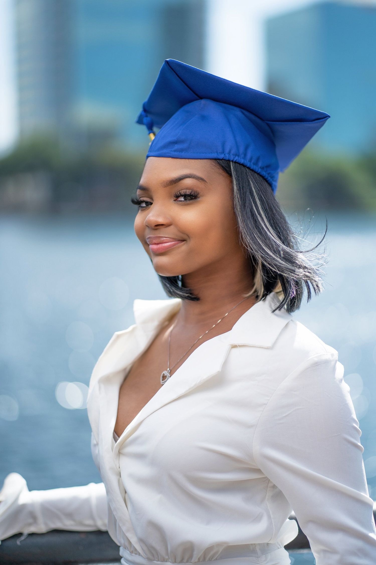 Graduate in blue cap and white shirt poses by waterfront with cityscape background.