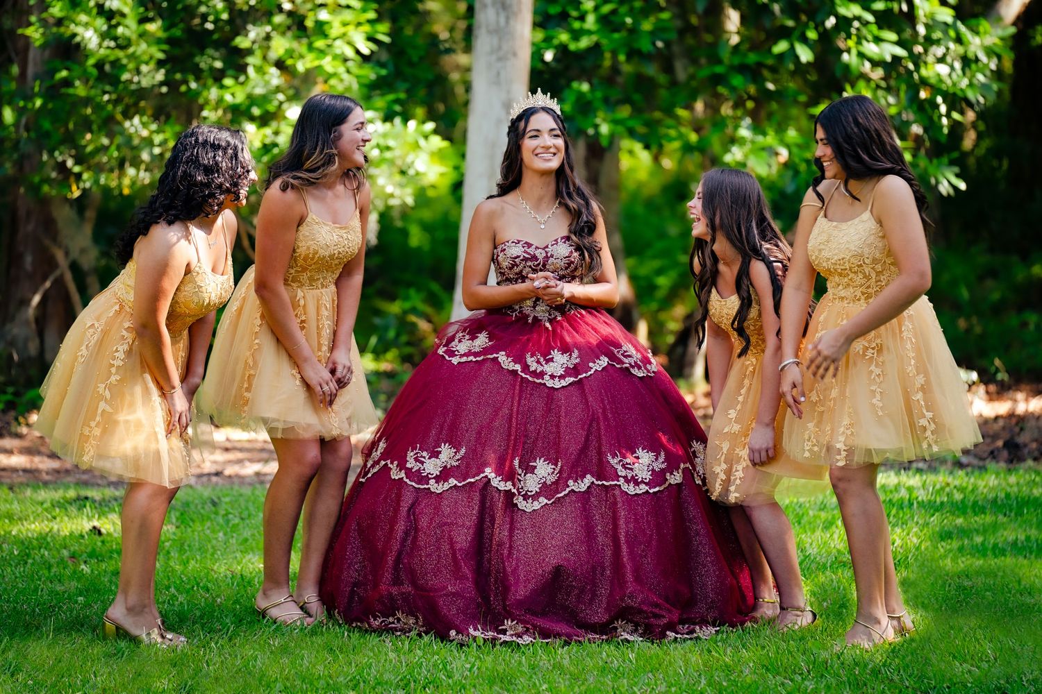 A quinceañera in a burgundy ball gown poses with four bridesmaids in gold dresses outdoors.