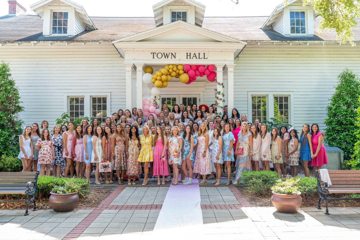 Large group of women in colorful dresses posing in front of a Town Hall building with balloon arch.