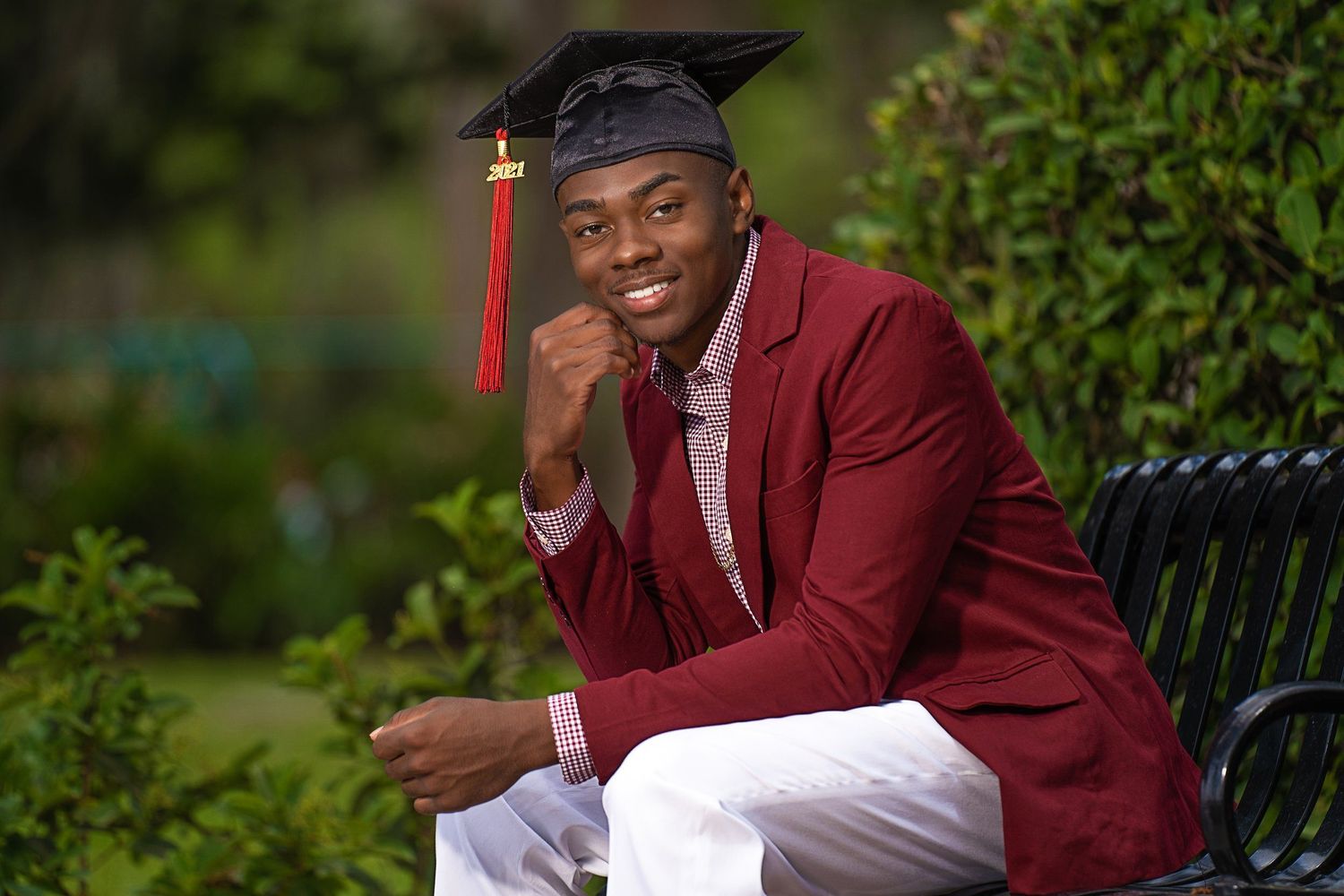 Smiling graduate in cap and maroon blazer with white pants sits on bench outdoors.