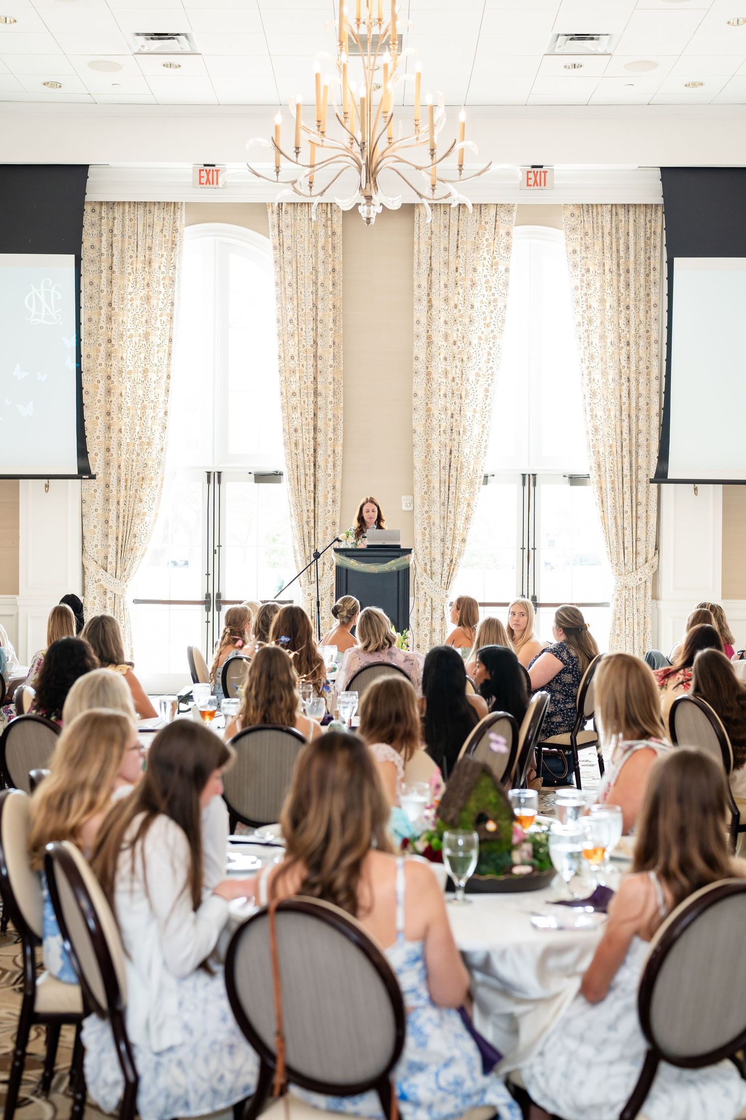 Women seated at elegant round tables listen to a speaker at a podium in a bright, chandelier-lit banquet hall.