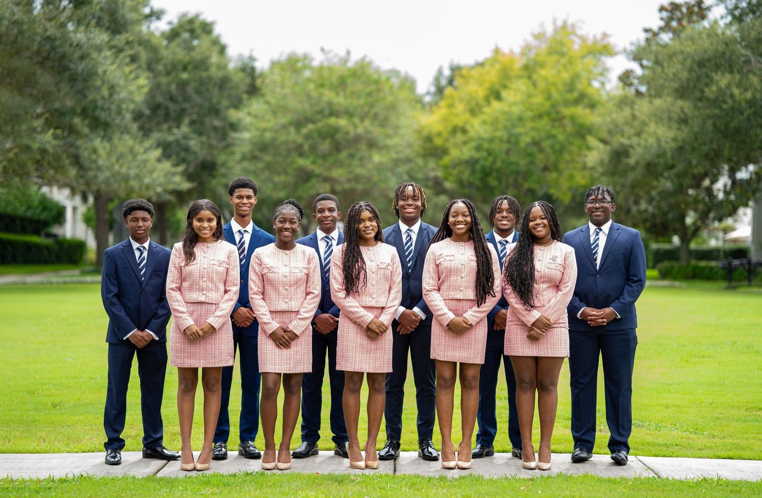 A group of ten young students in matching pink and navy formal attire posing outdoors on a green lawn.