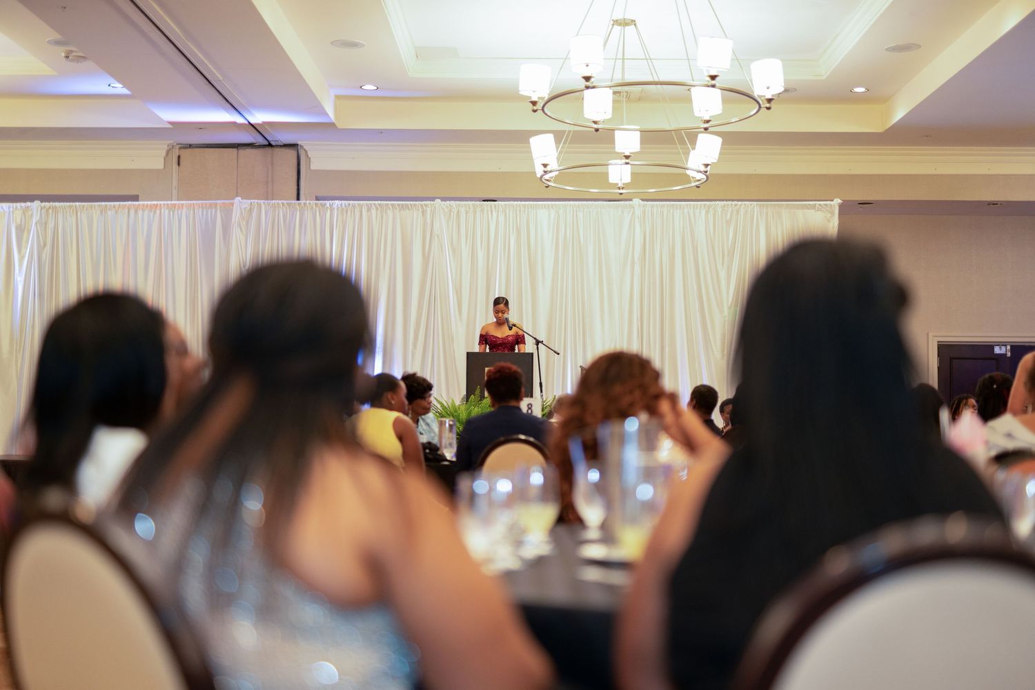 Woman speaking at podium during formal banquet event in elegant ballroom with chandelier and seated audience.