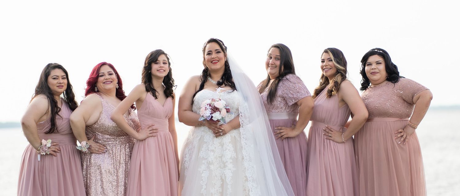 Bride in white lace gown and veil holds bouquet surrounded by six bridesmaids in dusty rose dresses outdoors.