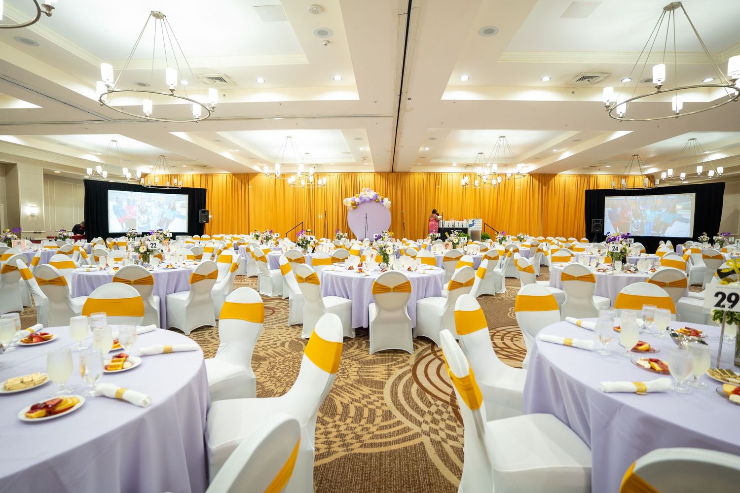Elegant banquet hall with round tables, white linens, yellow chair sashes, and projection screens for a formal event.