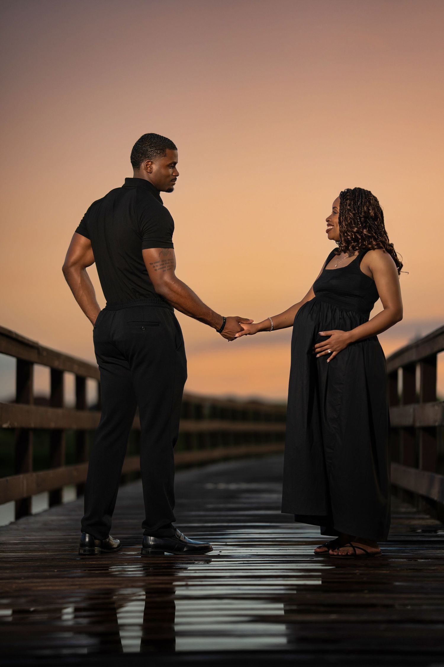 Expecting couple in black outfits hold hands on a wooden pier at sunset, woman cradling baby bump.