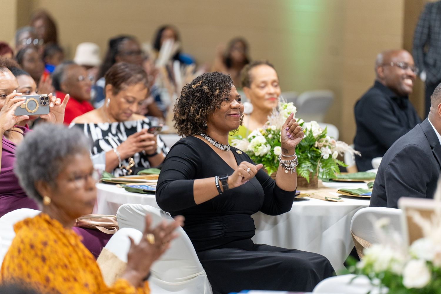 Guests clapping and celebrating at a formal banquet event, with floral centerpieces on white-clothed tables.