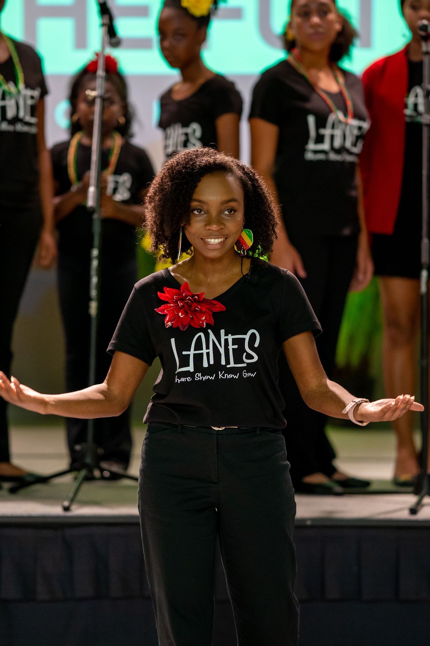 Young woman in LANES black t-shirt performing on stage with choir group in matching shirts behind her.