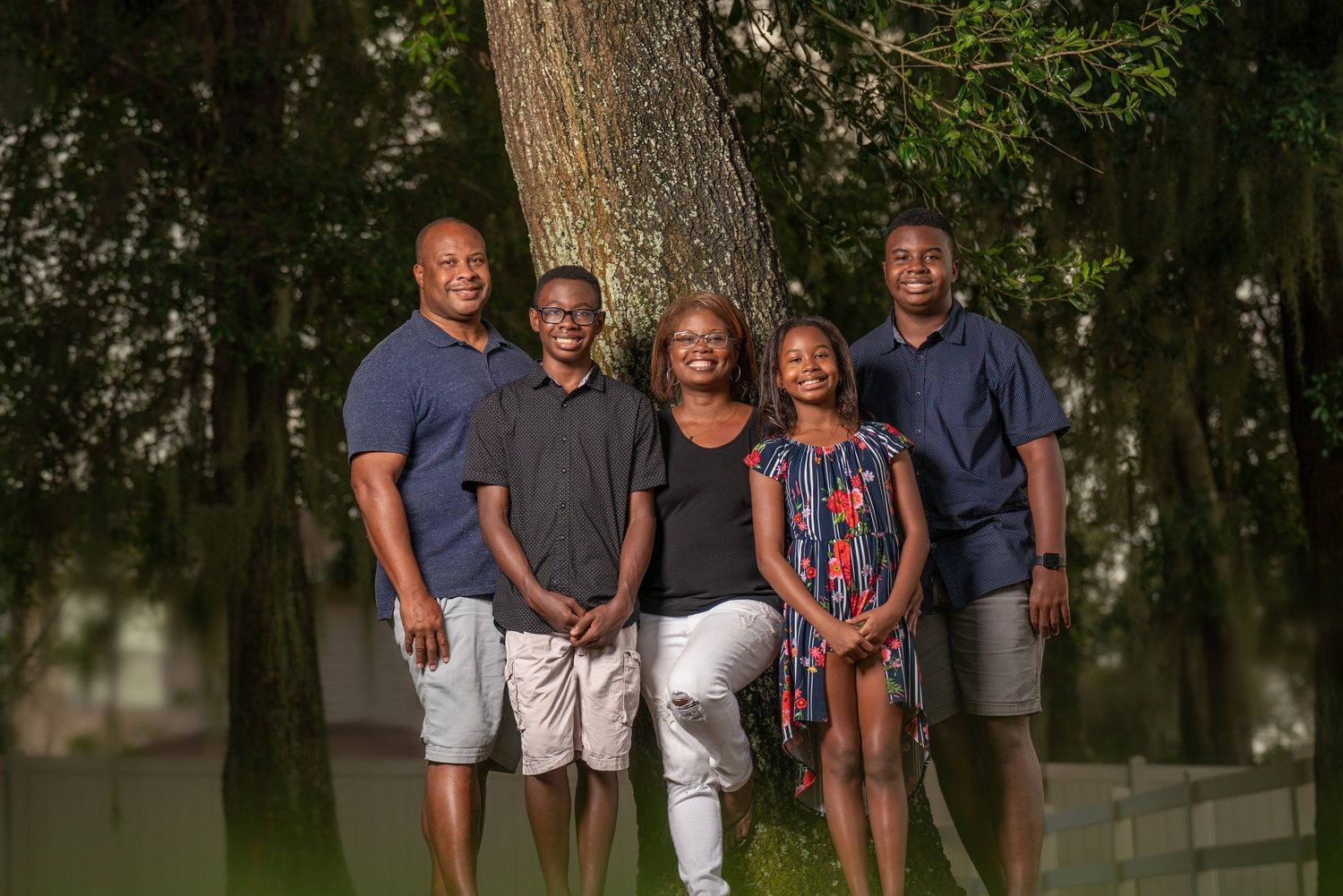 A joyful family of five poses together outdoors near a large tree, smiling in casual attire on a sunny day.
