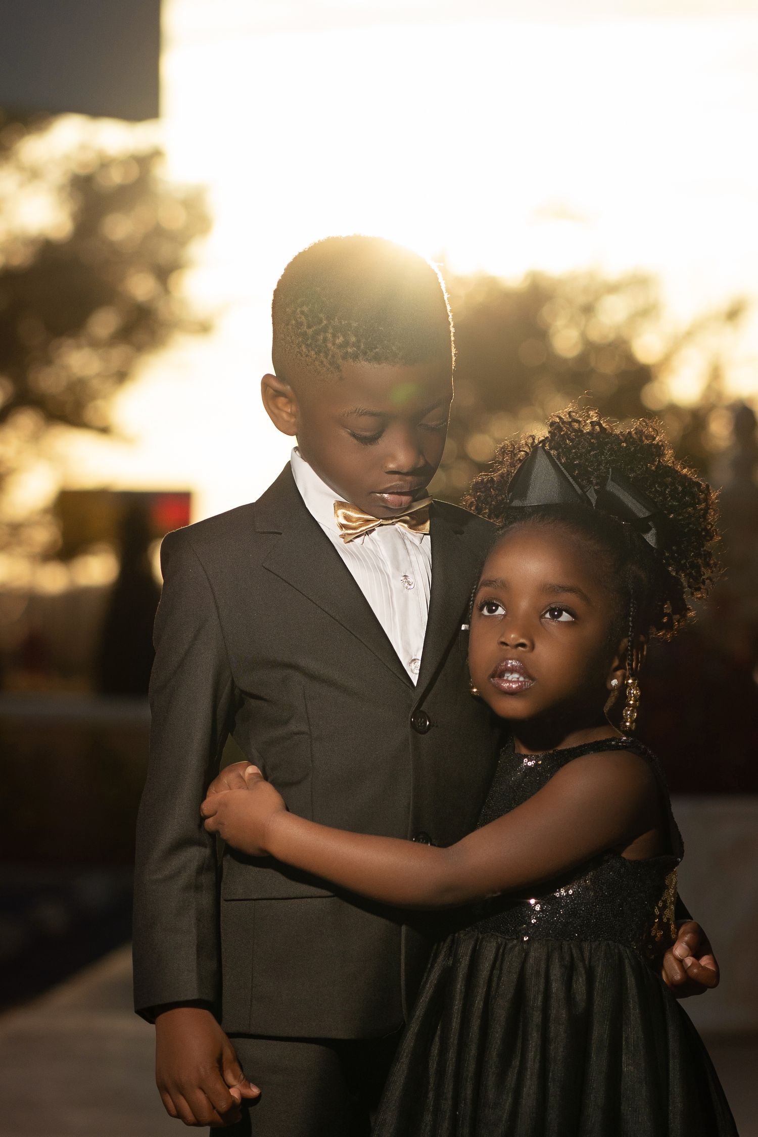 Two young Black children dressed in formal attire embrace during a golden sunset outdoor photoshoot.