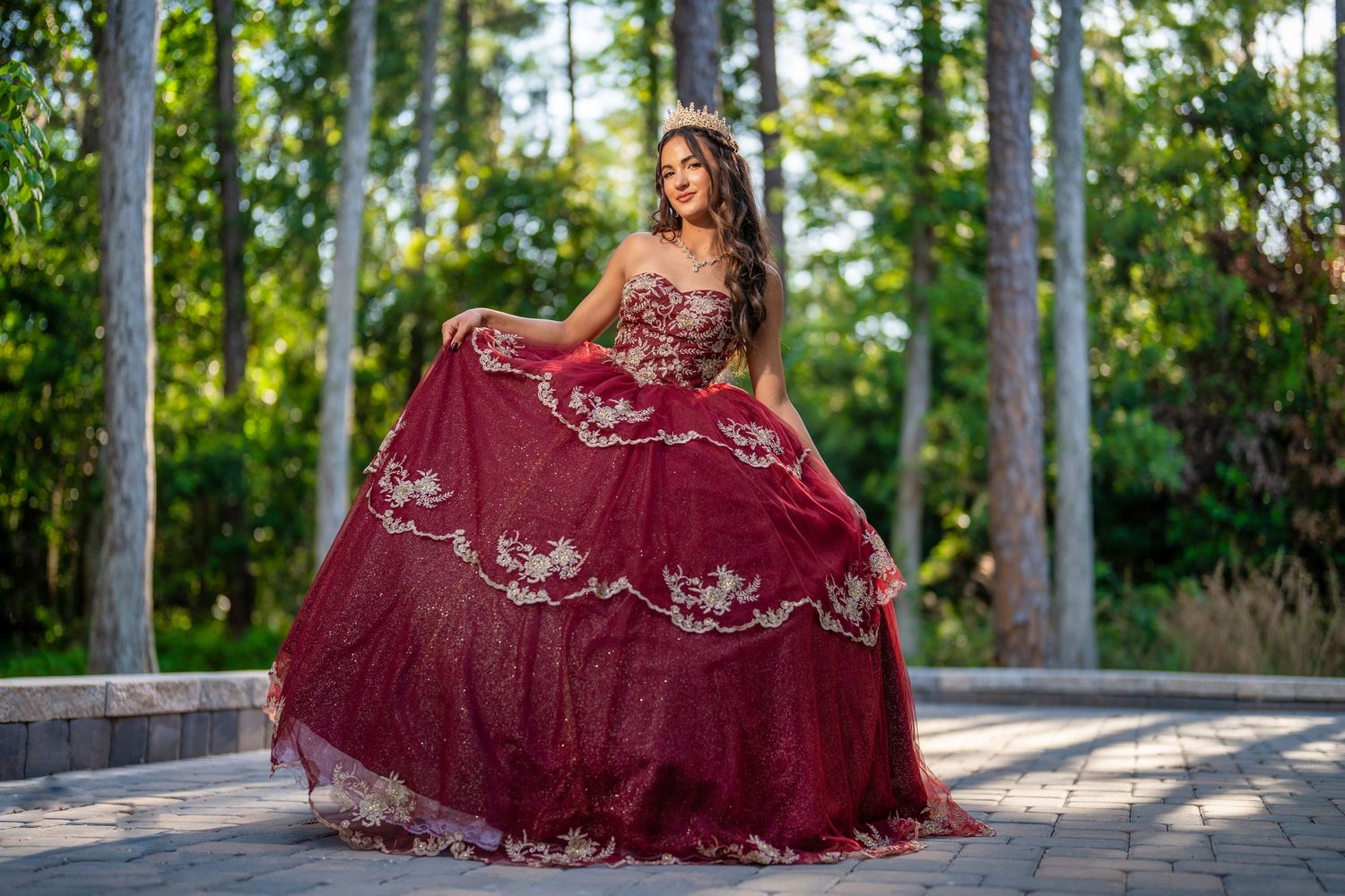 A burgundy ball gown with silver embroidery photographed in a sunlit garden setting.