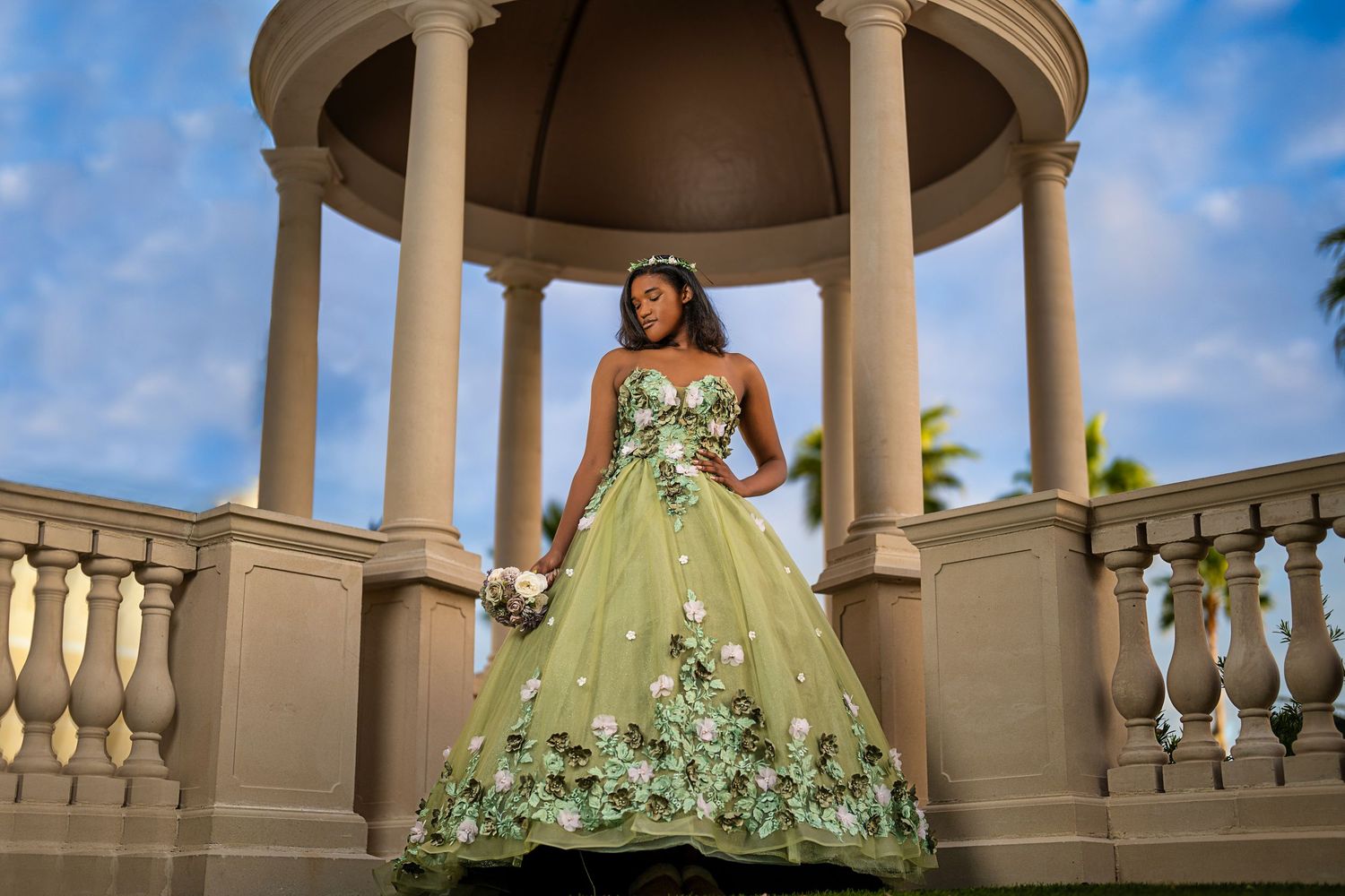 Young woman in green floral quinceañera ball gown poses in elegant gazebo with blue sky backdrop.