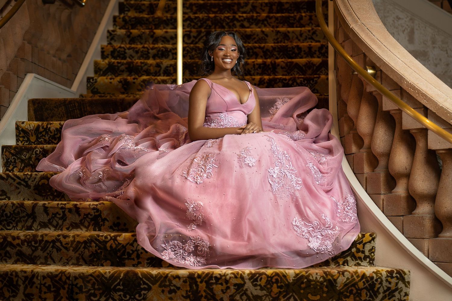 A young Black woman in a stunning pink ball gown with lace details poses gracefully on ornate golden stairs.