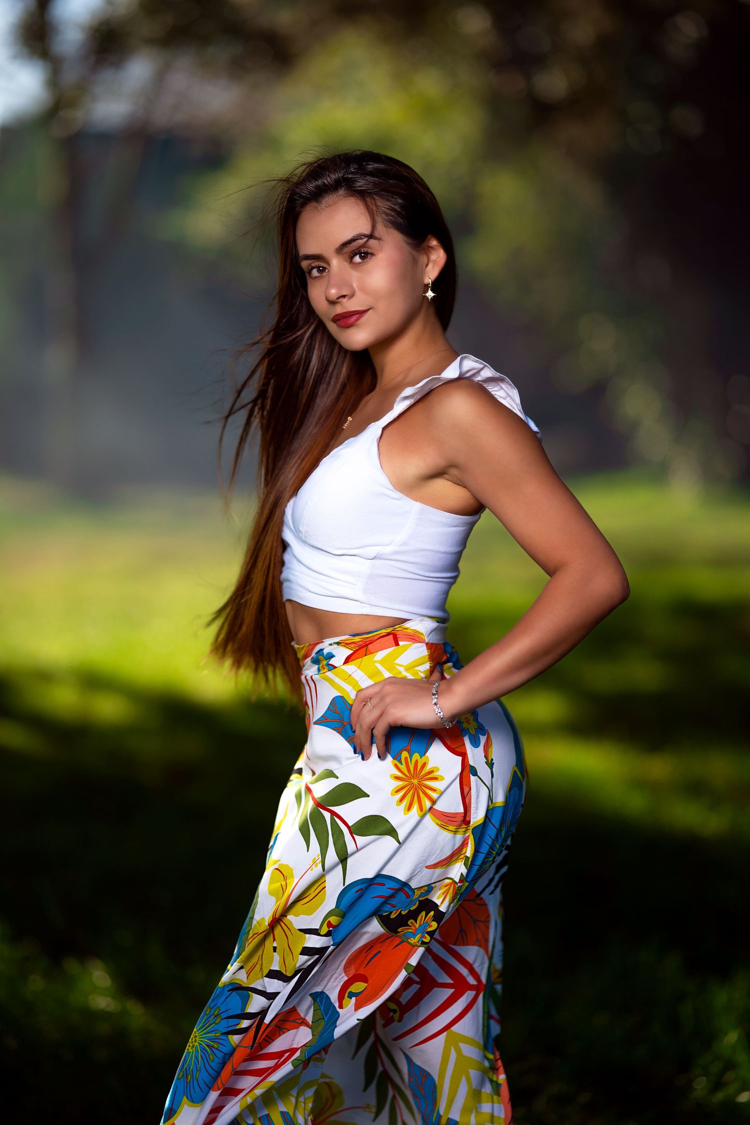 Young woman in white crop top and colorful floral skirt posing outdoors in a sunlit green park setting.