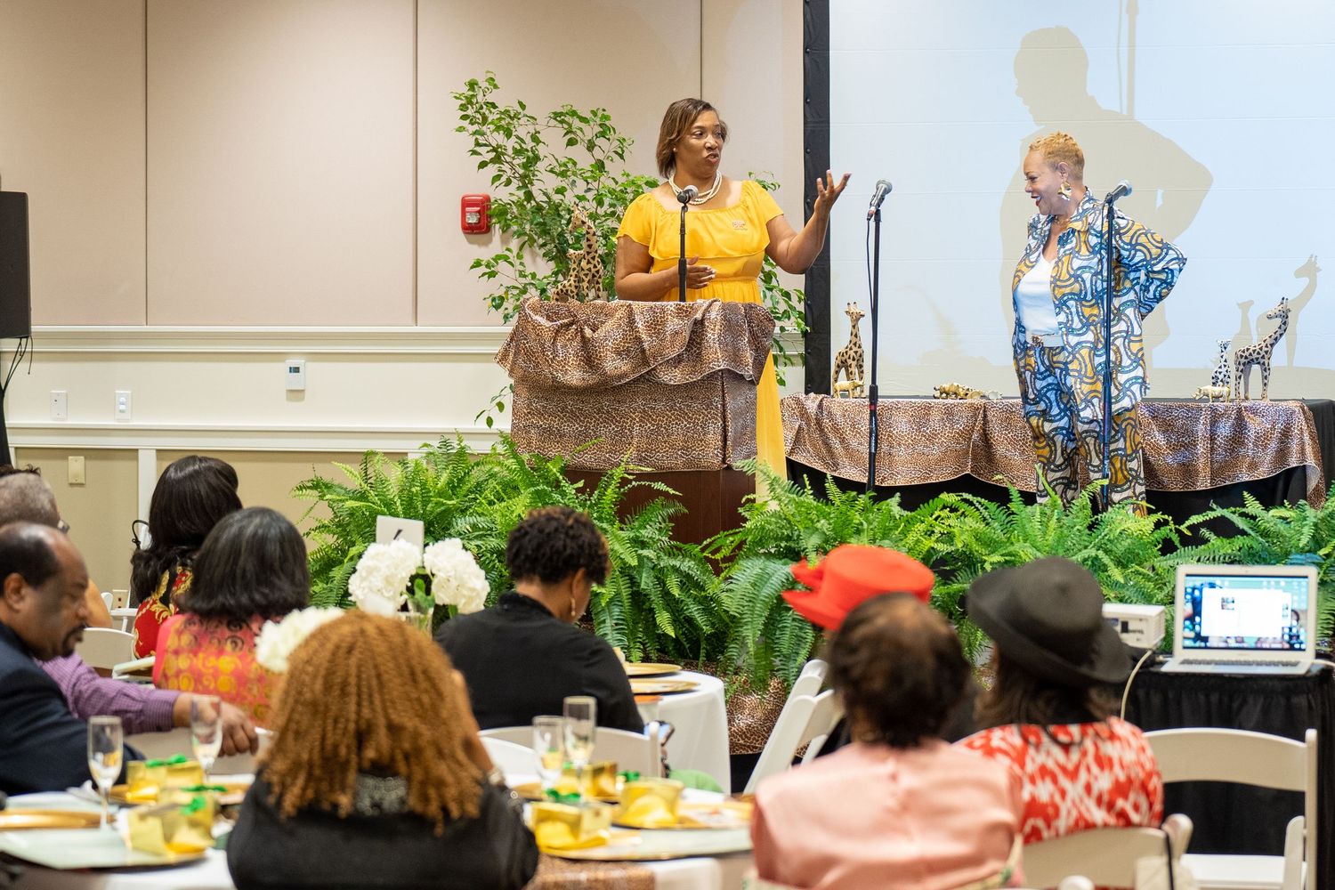 Two women presenting on stage at a formal banquet event with guests seated at decorated tables.