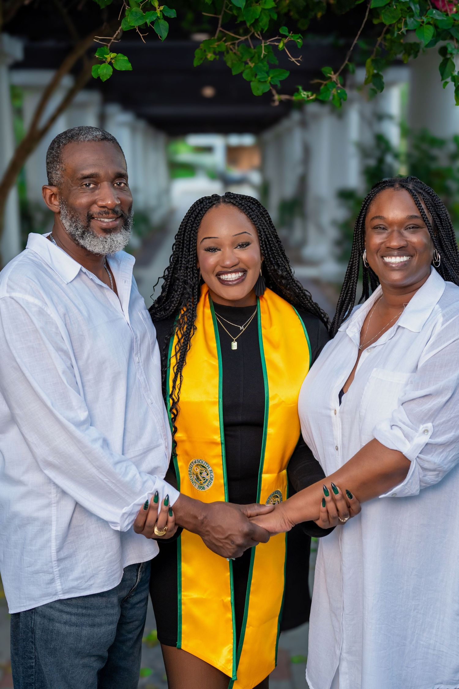 A graduate in yellow and green stole poses with two proud family members, all smiling and holding hands outdoors.